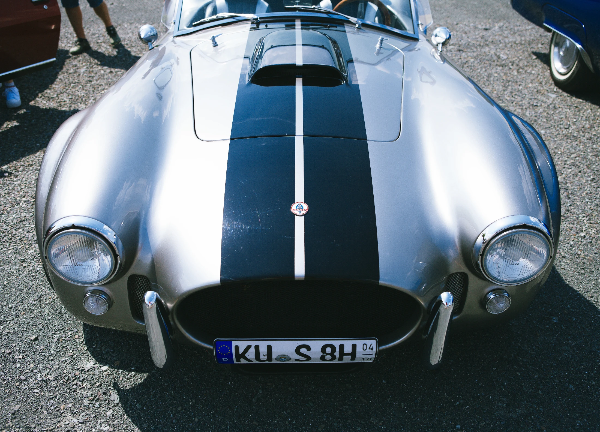 Front view of a silver AC Cobra-style roadster with dark racing stripes and round headlights