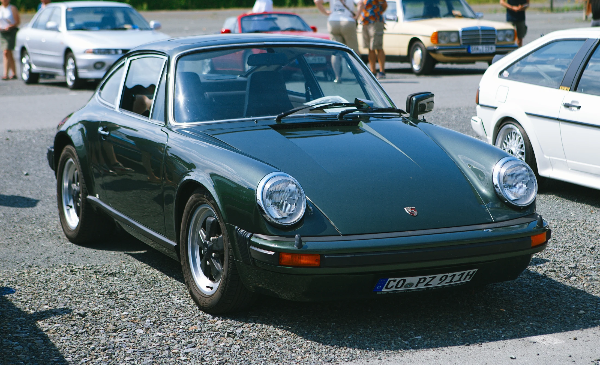 Dark green classic Porsche 911 with round headlights and black wheels, viewed from the front three-quarter angle